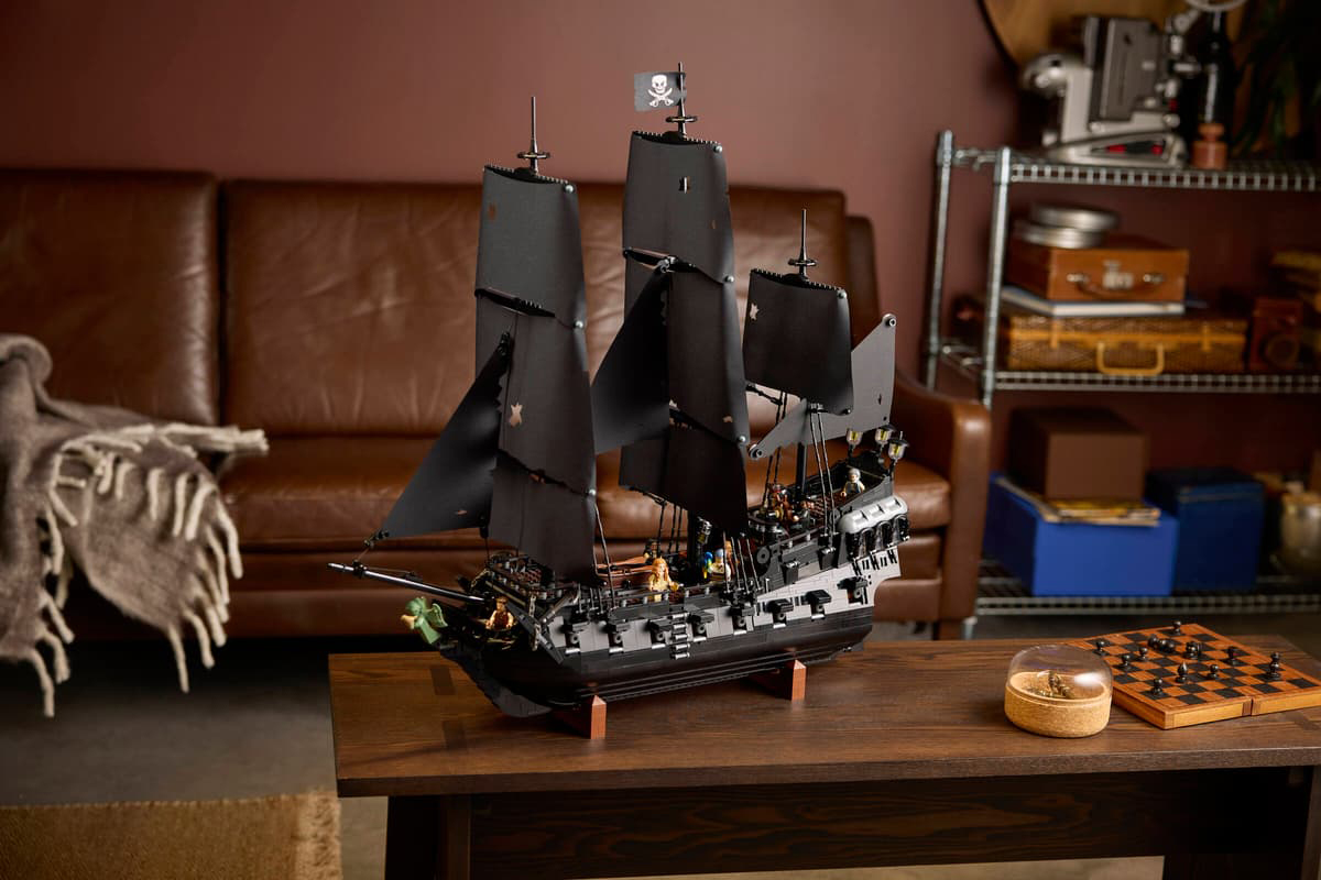 Czarny statek piracki LEGO Icons 10365 wyeksponowany w przytulnym pokoju na drewnianym stole obok szachownicy. Konstrukcja z postrzępionymi żaglami i flagą z czaszką stanowi główny element ujęcia, a w tle widać skórzaną kanapę oraz regał.
