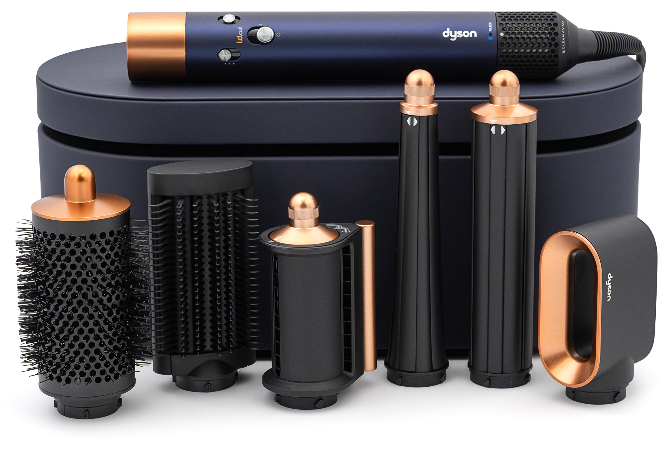 Zestaw do stylizacji włosów Dyson Airwrap i.d. w kolorze błękitu pruskiego z miedzianymi akcentami. W centralnej części widoczny jest główny korpus urządzenia z przyciskami sterującymi. Pod nim umieszczone jest granatowe etui, przed którym ustawiono sześć wymiennych końcówek: szczotkę okrągłą, szczotkę do wygładzania, nasadkę do stylizacji, dwie końcówki do kręcenia włosów oraz nasadkę wygładzającą typu Coanda. Wszystkie elementy zestawu zostały zaprezentowane na czystym, białym tle.