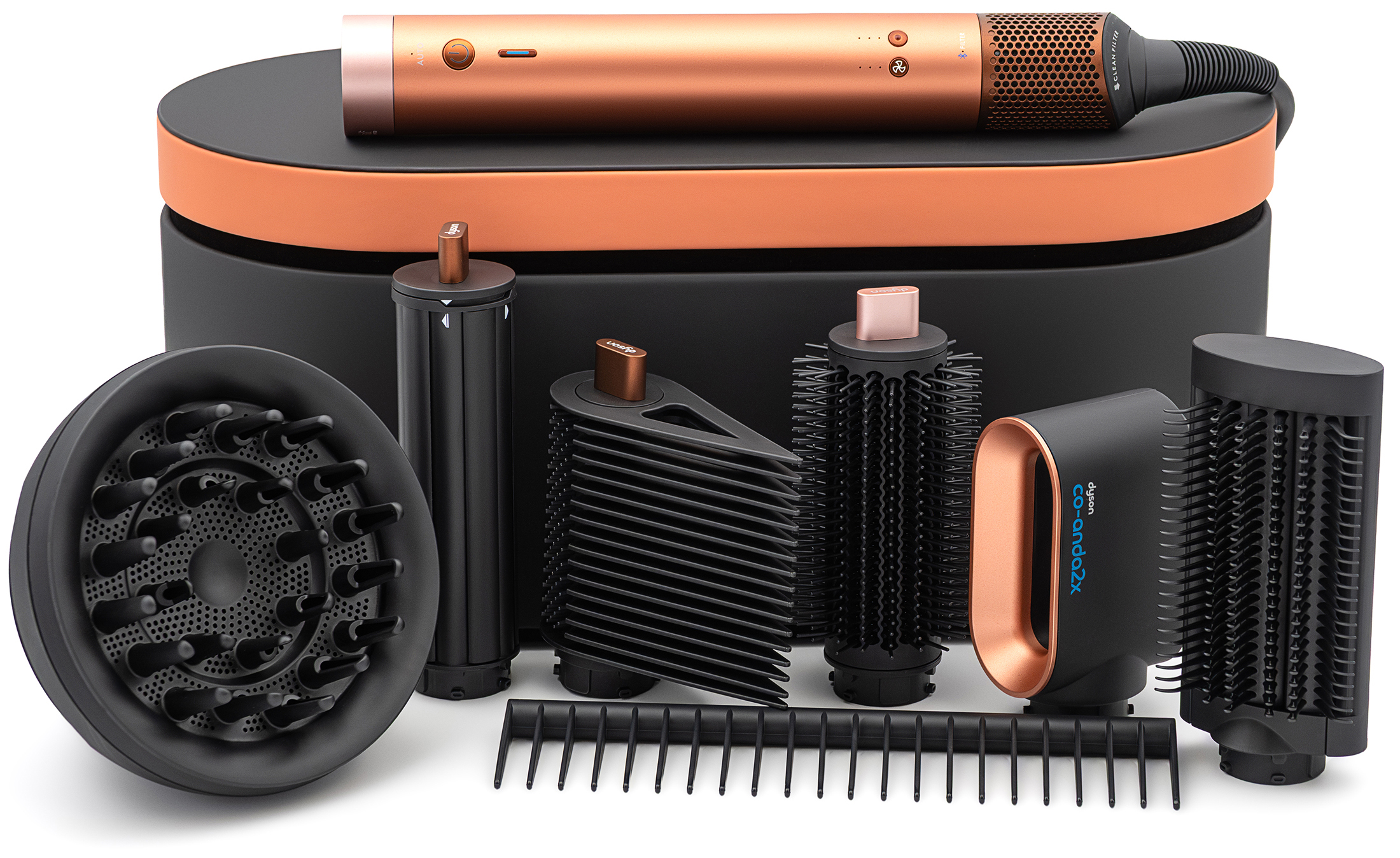 Zestaw Dyson Airwrap Co-anda2x Curly+Coily w odcieniu bursztynowego jedwabiu, zaprezentowany na białym tle. W centralnej części widoczne jest czarne, owalne etui z miedzianą pokrywą, na którym spoczywa miedziany korpus urządzenia. Przed etui ułożono w siedem akcesoriów: dyfuzor, nasadkę cylindryczną, końcówkę do wygładzania, okrągłą szczotkę, nasadkę Co-anda z oznaczeniem „co-anda2x”, szczotkę wygładzającą oraz czarny grzebień.