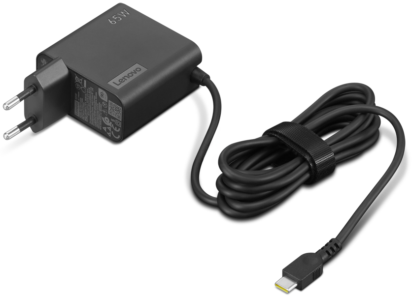 Czarny zasilacz Lenovo 65W USB-C na białym tle, widok pod kątem z góry. Matowa ładowarka posiada widoczne oznaczenie 65W, logo LENOVO oraz naklejkę z danymi technicznymi i certyfikatami obok wtyczki sieciowej. Czarny przewód zasilający jest zwinięty, spięty rzepem i zakończony wtyczką USB-C.