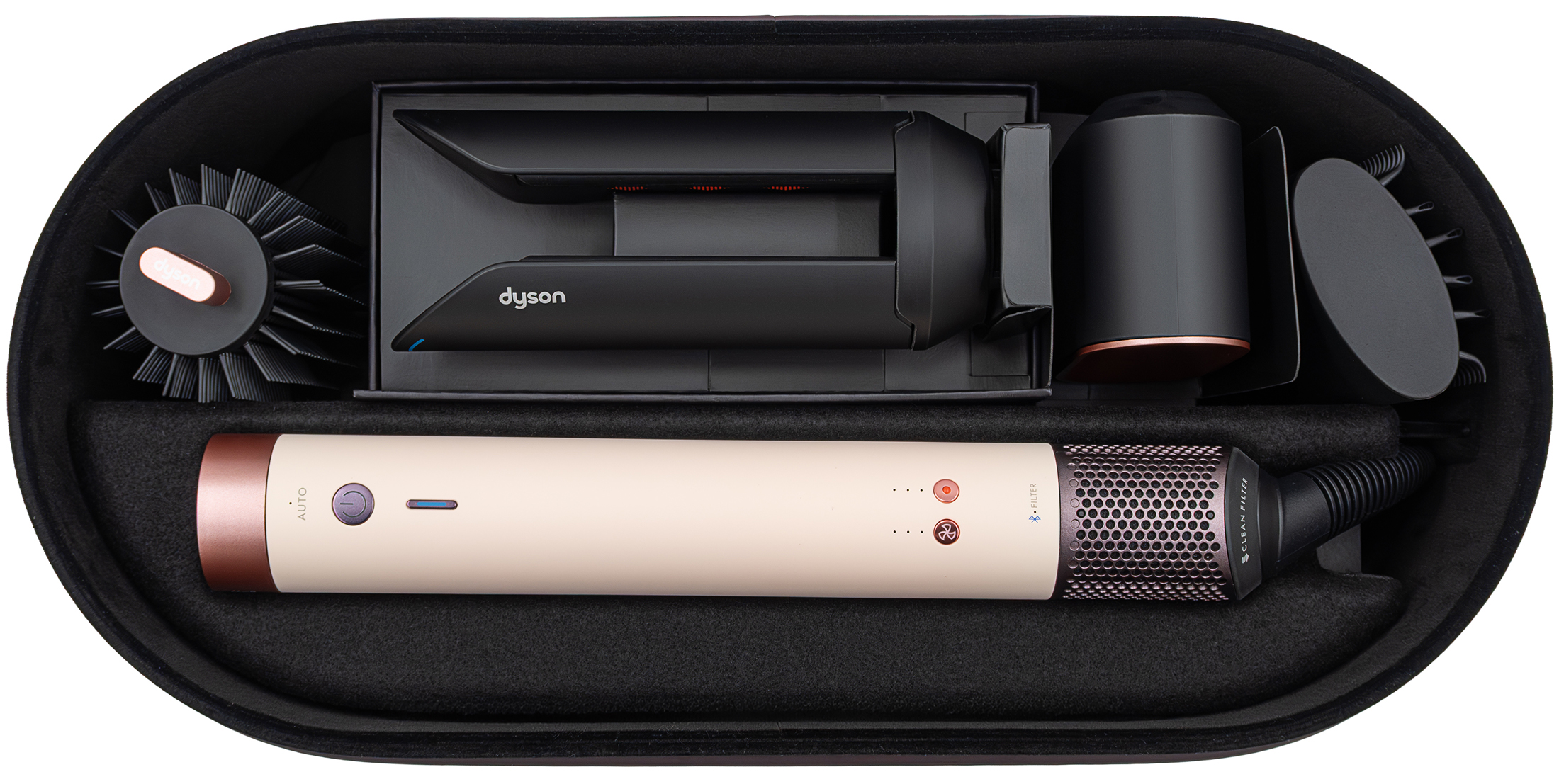 Widok z góry na otwarte etui multistylera Dyson Airwrap Co-anda2x. W środku znajduje się jasnoróżowa rączka oraz cztery czarne końcówki do stylizacji, starannie ułożone w dopasowanych przegrodach. Wnętrze etui jest czarne, co kontrastuje z jasnymi elementami urządzenia.