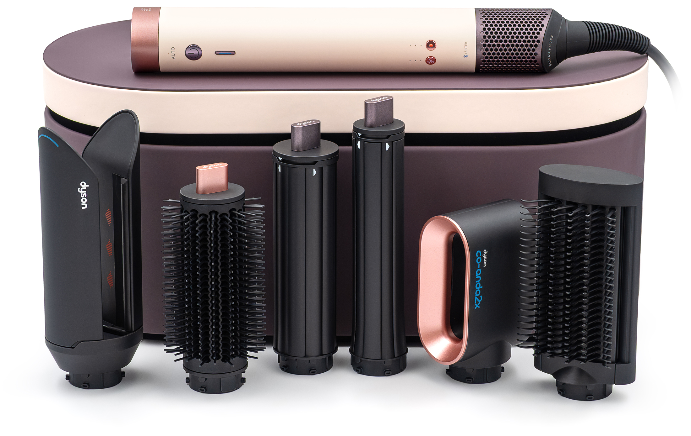 Zestaw Dyson Airwrap Co-anda2x Straight+Wavy z fioletowym etui i sześcioma czarnymi końcówkami do stylizacji. Główna rączka urządzenia w kolorze różowego złota leży na zamkniętym kuferku. Całość ukazana w widoku z przodu na białym tle.