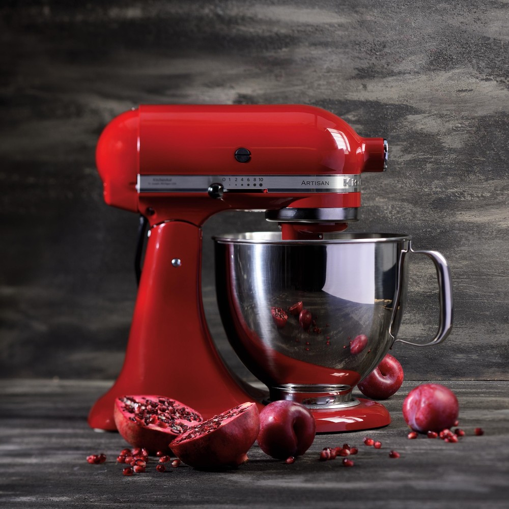 Czerwony robot kuchenny KitchenAid 5KSM125EER na ciemnym blacie, z granatem i śliwkami leżącymi z przodu, na tle drewnianej ściany.