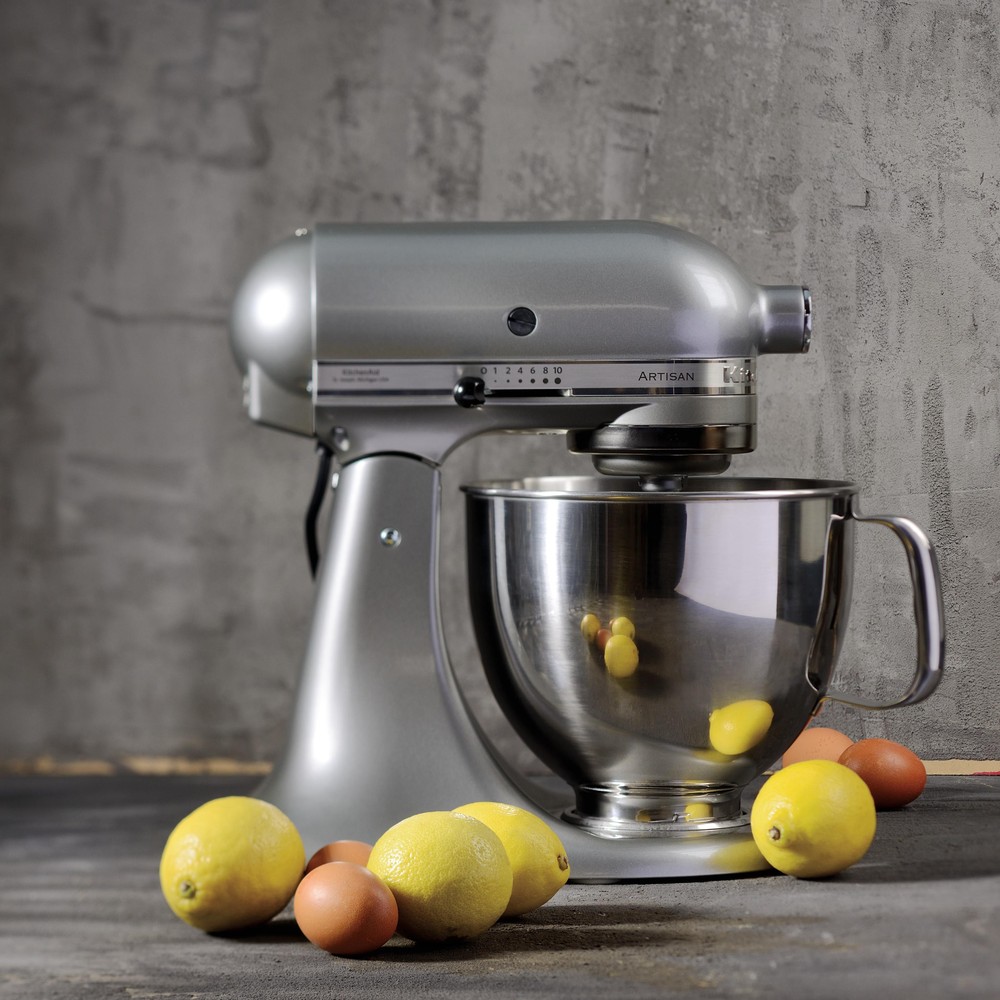 Srebrny robot kuchenny KitchenAid 5KSM125ECU na ciemnym blacie, z cytrynami i jajkami leżącymi z przodu, na tle betonowej ściany.