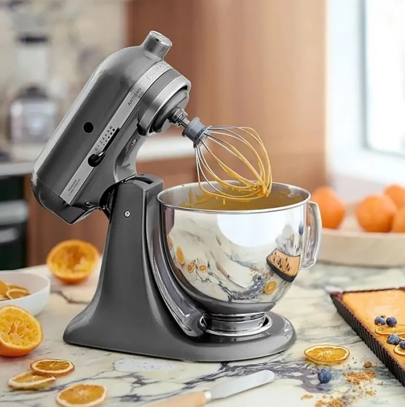 Robot kuchenny KitchenAid Artisan w kolorze srebrnym pracuje na marmurowym blacie, ubijając pomarańczowy krem w metalowej misie za pomocą drucianej trzepaczki. Wokół maszyny leżą połówki i plasterki pomarańczy, a obok widoczny jest fragment tarty.