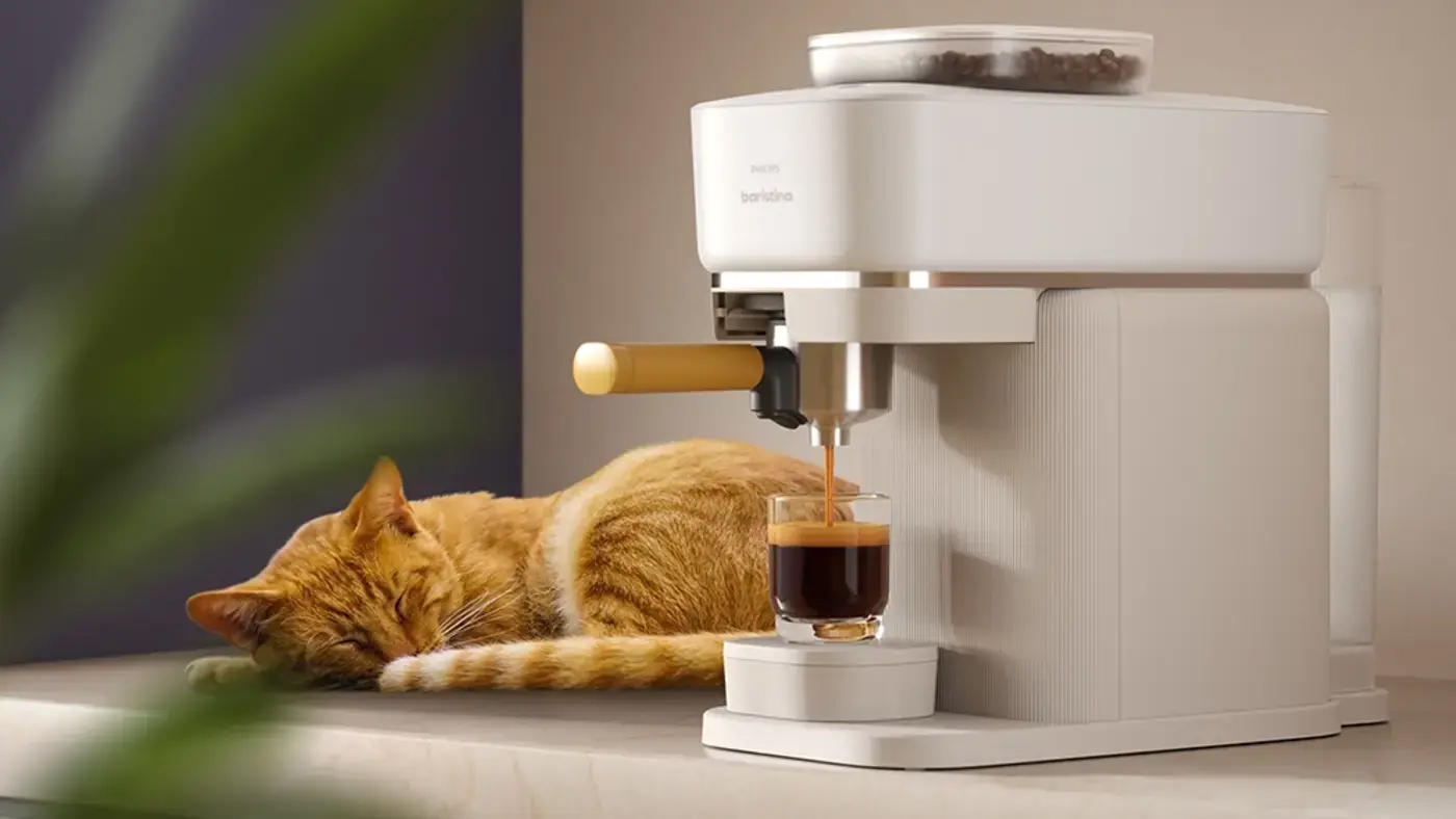 Biały ekspres Philips Baristina BAR300/03 w trakcie nalewania kawy do szklanki na blacie kuchennym. Obok urządzenia śpi rudy kot. Ekspres wyróżnia się żółtym uchwytem kolby oraz przezroczystym pojemnikiem na ziarna kawy u góry.