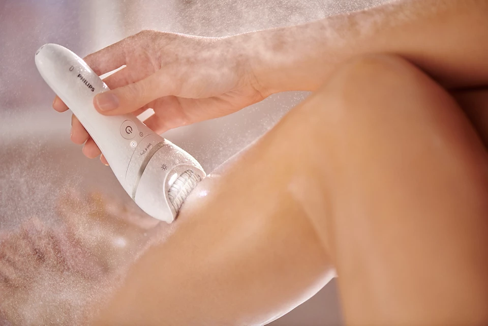 Dłoń używa depilatora Philips BRE770/92 do golenia nogi na mokro. W powietrzu widać drobne kropelki wody.