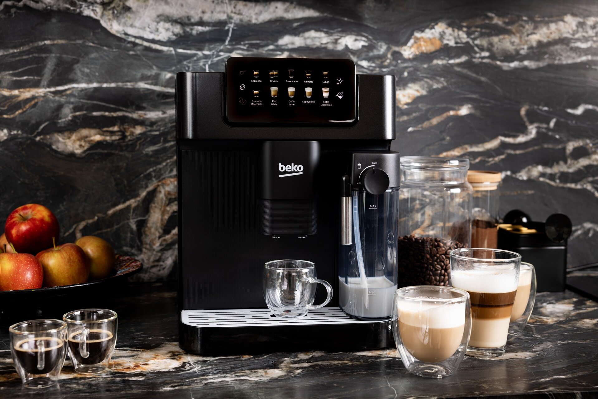 Ekspres Beko CaffeExperto CEG7304B w kolorze czarnym, w aranżacji kuchennej na marmurowym blacie. Obok ekspresu stoją dwie szklanki z kawami mlecznymi i słoik z ziarnami kawy. Widoczny jest podświetlany panel i pojemnik na mleko.