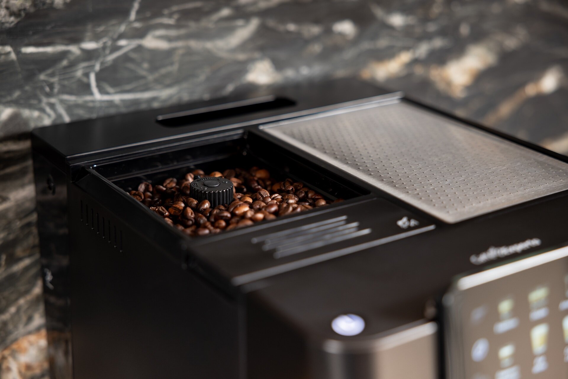 Zbliżenie na otwarty zbiornik na ziarna czarnego ekspresu Beko CEG7304B. Widoczne są świeże ziarna kawy i pokrętło regulacji mielenia. Z boku znajduje się srebrna kratka.