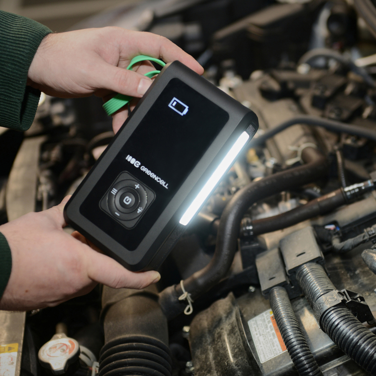 Jump Starter Green Cell PowerBoost Air 3w1 trzymany nad silnikiem samochodu. Urządzenie ma włączoną boczną latarkę LED, która oświetla wnętrze maski. Na ekranie widoczna jest ikona stanu naładowania, a zielony pasek na nadgarstek ułatwia bezpieczne trzymanie urządzenia podczas pracy przy aucie.