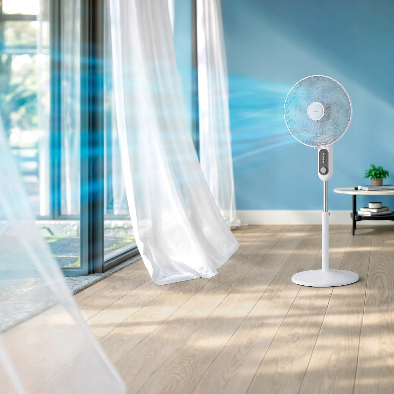 Biały wentylator stojący Philips Seria 1000 CX1220/00 emituje błękitny strumień powietrza, poruszający białymi zasłonami w salonie. Urządzenie stoi na drewnianej podłodze obok stolika z zieloną rośliną.