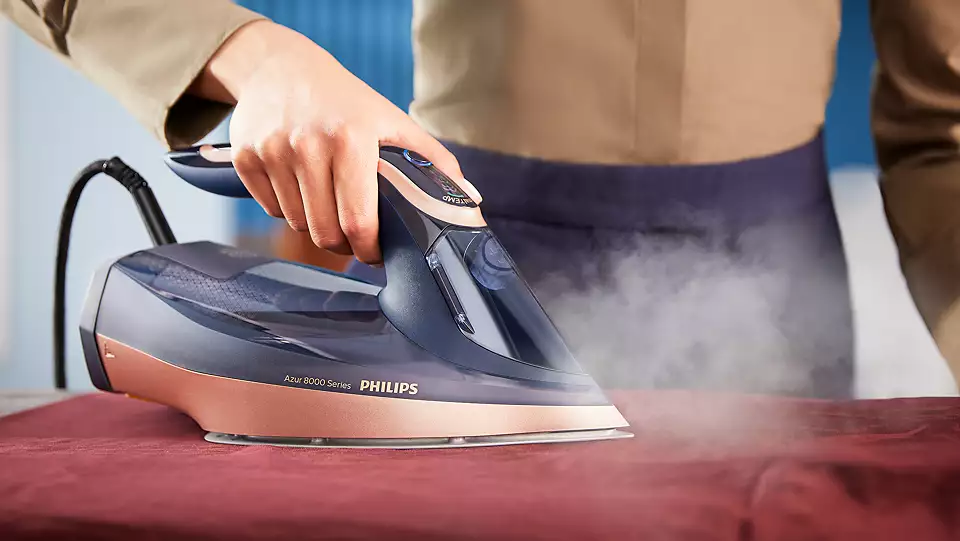 Kobieta prasuje bordowy materiał żelazkiem parowym Philips DST8050/20. Z urządzenia wydobywa się duży strumień pary.