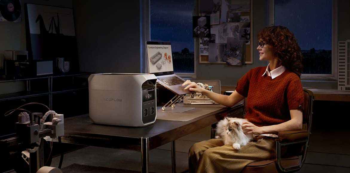 Stacja zasilania EcoFlow Delta 3 Classic zasila laptopa na biurku, przy którym pracuje kobieta z kotem na kolanach. Scena dzieje się nocą w przytulnym, domowym biurze. Urządzenie z włączonym wyświetlaczem stoi wyeksponowane na pierwszym planie blatu.