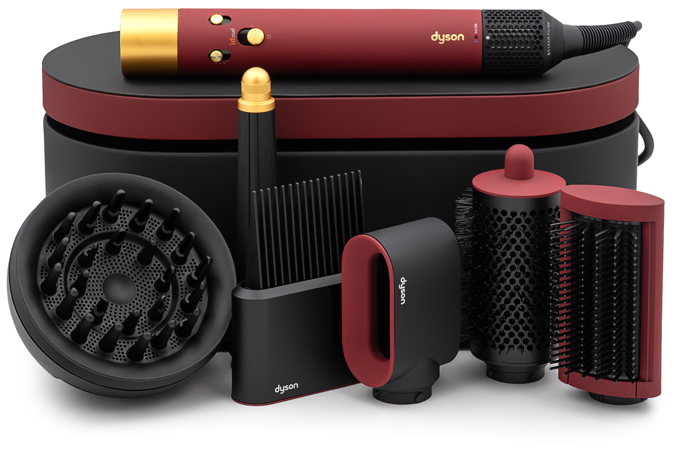 Zestaw stylizacyjny Dyson Airwrap i.d. Curly+Coily w kolorze aksamitnej czerwieni i złota. Urządzenie leży na czarnym etui, w otoczeniu zestawu wymiennych końcówek, w tym dyfuzora i szczotek. Całość zaprezentowana na białym tle.