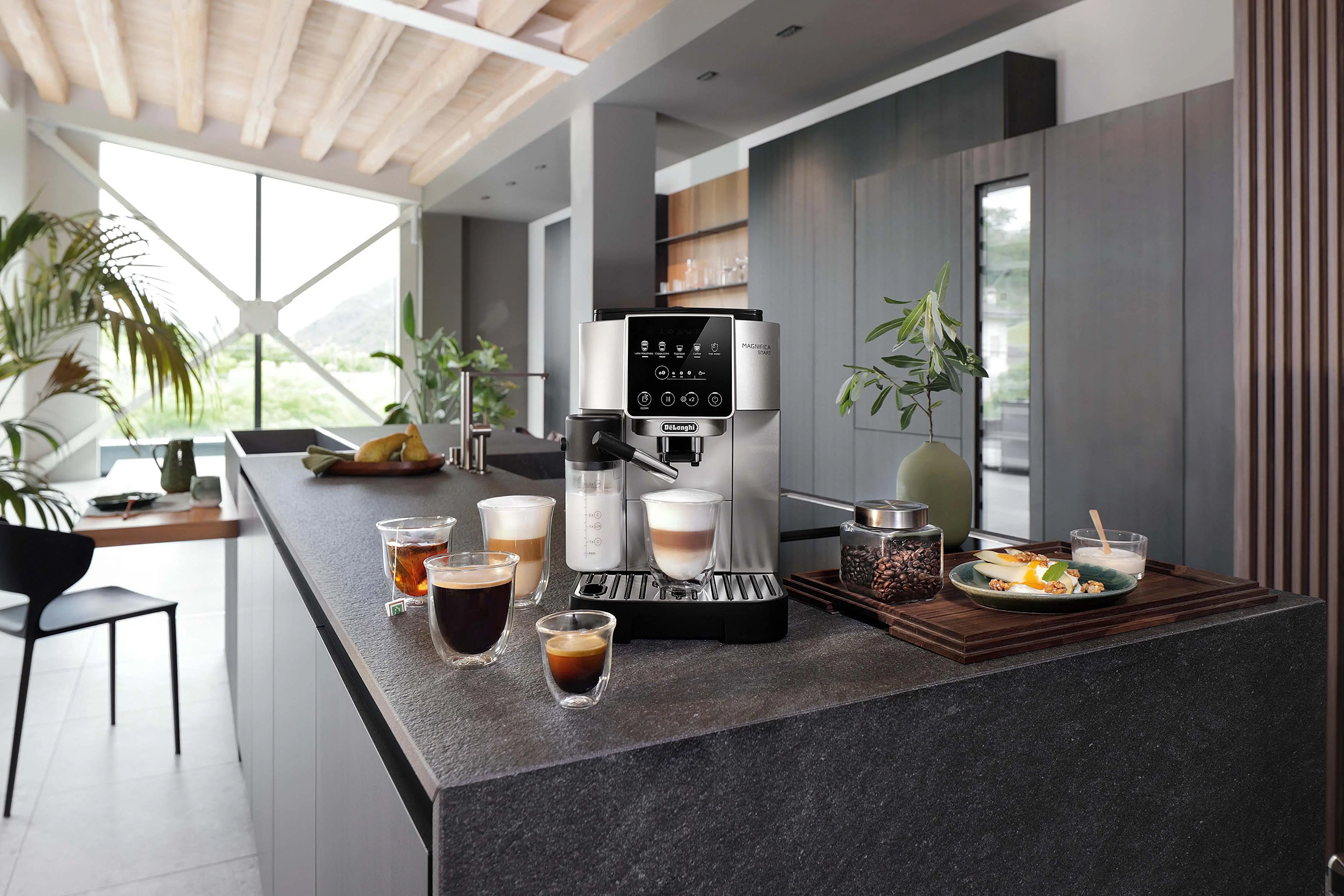 Ekspres do kawy DeLonghi Magnifica Start ECAM220.80.SB na blacie wyspy kuchennej, obok stoją szklanki z różnymi rodzajami gotowej kawy. W tle nowoczesna kuchnia z oknami i widokiem na zewnątrz.