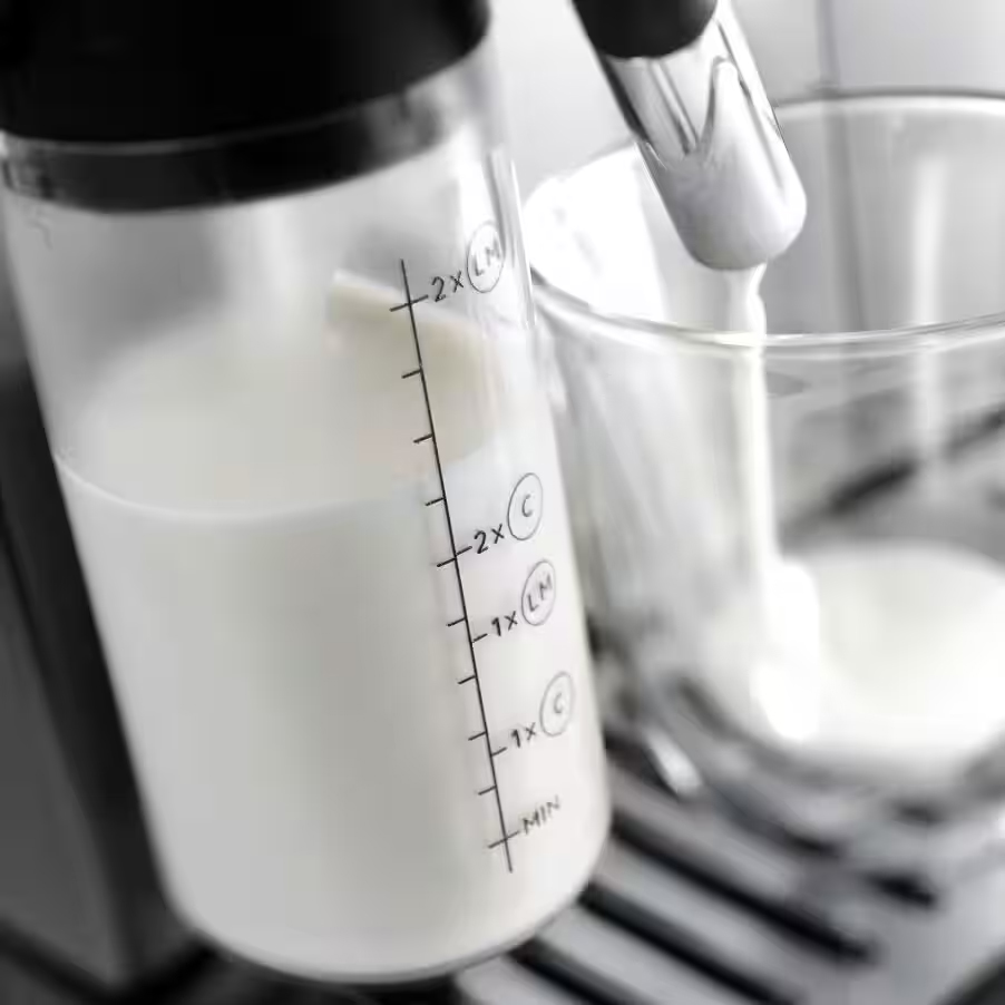 Zbliżenie na ekspres DeLonghi, ukazujące proces wlewania spienionego mleka do szklanki z przezroczystego pojemnika.