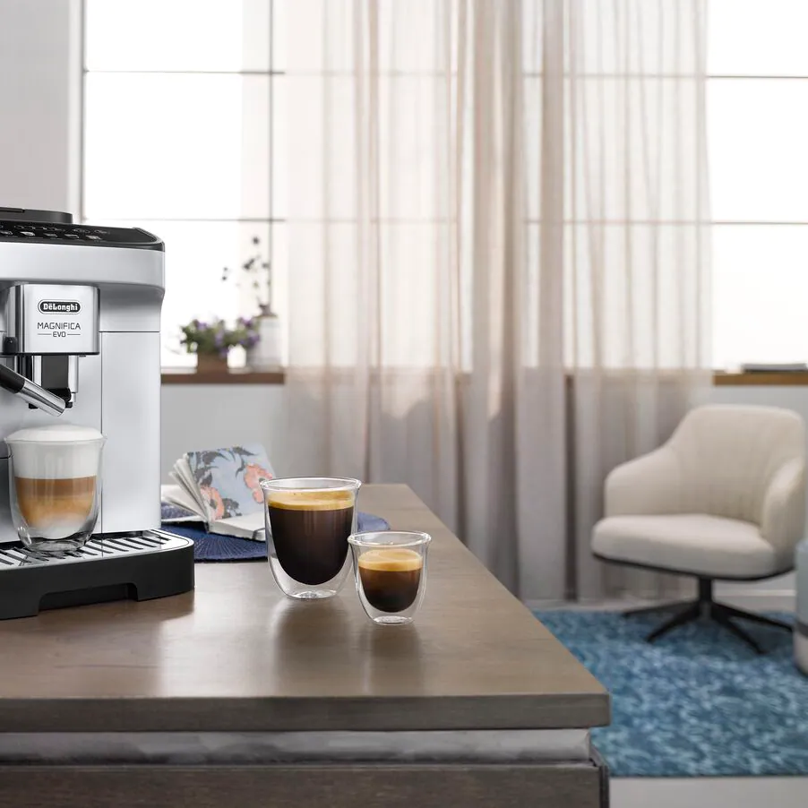 Ekspres ciśnieniowy DeLonghi ECAM290.61.SB na stole w salonie, przygotowujący kawę, obok dwie szklanki z kawą.