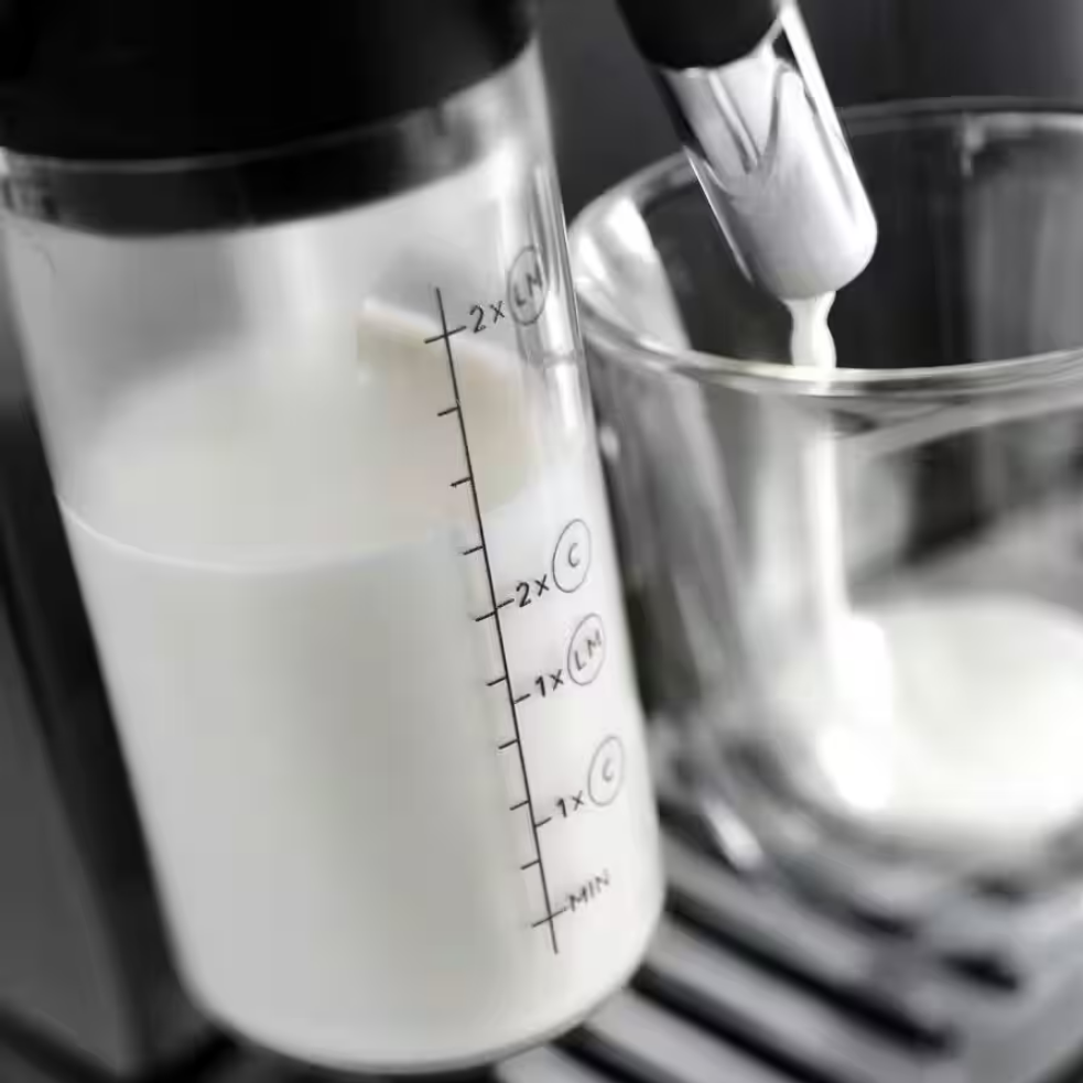 Zbliżenie na ekspres DeLonghi, ukazujące proces wlewania spienionego mleka do szklanki z przezroczystego pojemnika.