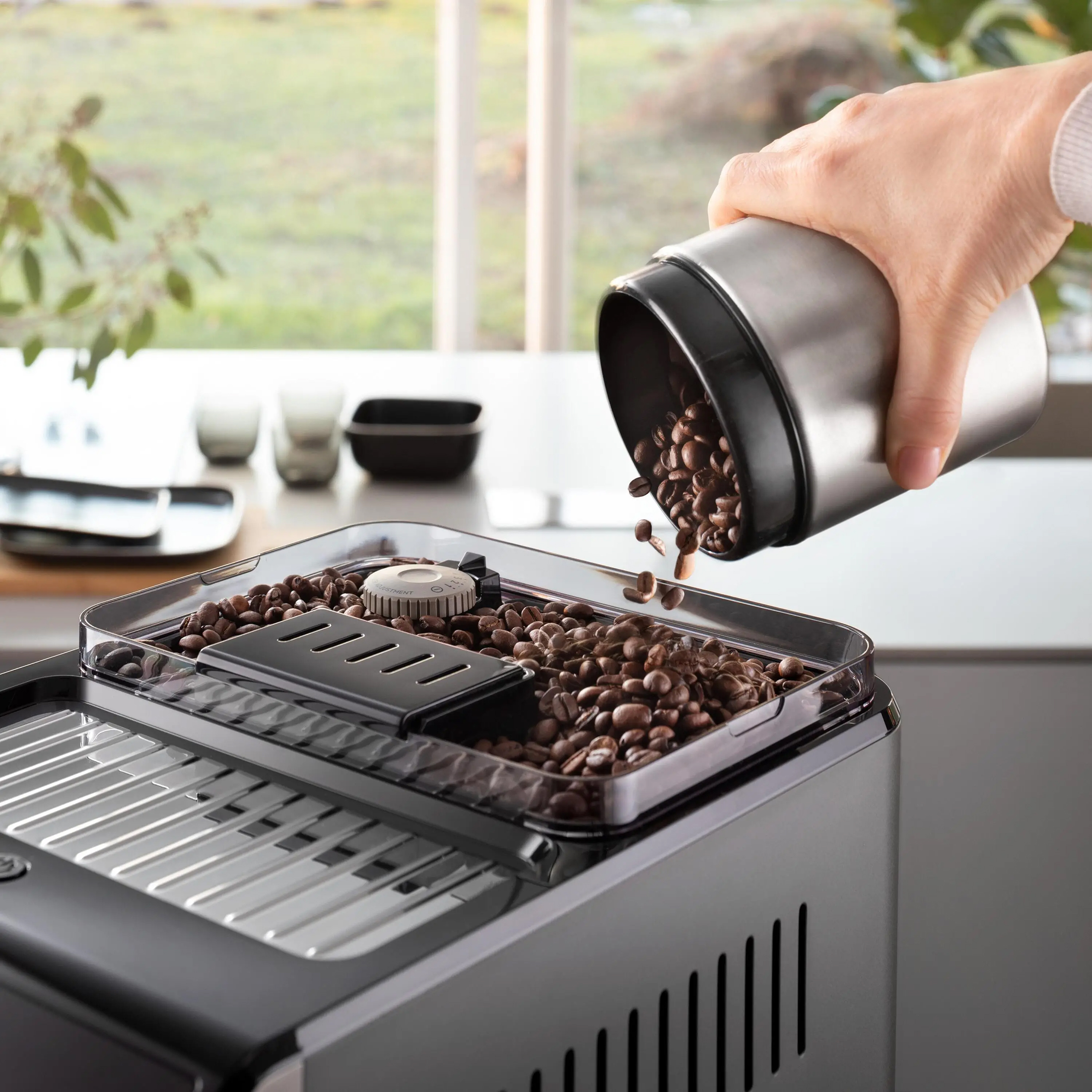 Ręka wsypuje ziarna kawy z metalowego pojemnika do otwartego zasobnika czarnego ekspresu DeLonghi Eletta Explore. Zdjęcie wykonane w jasnym wnętrzu kuchennym z dużym oknem.