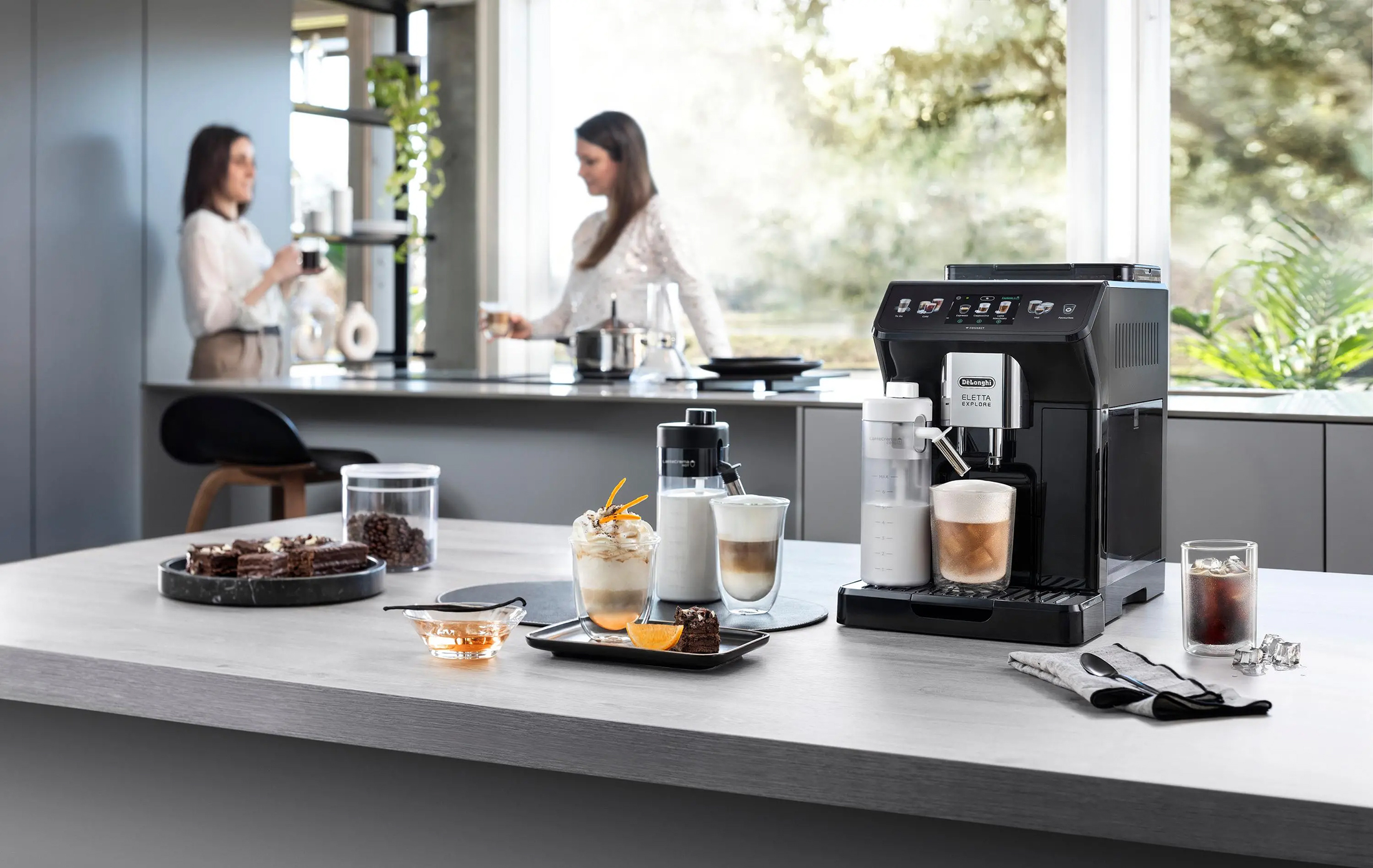 Ekspres DeLonghi Eletta Explore na szarym blacie kuchennym, otoczony akcesoriami, gotowymi kawami i deserami. W tle, przy oknie, widoczne są dwie kobiety w nowoczesnej kuchni.