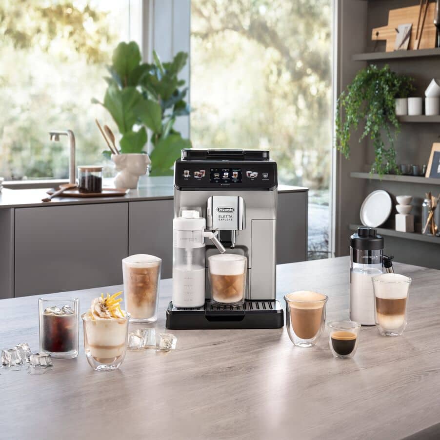 Srebrny ekspres DeLonghi Eletta Explore w jasnej kuchni, otoczony bogatym wyborem kaw gorących i mrożonych. Na blacie widoczne są obie karafki systemu LatteCrema oraz gotowe napoje w szklankach.