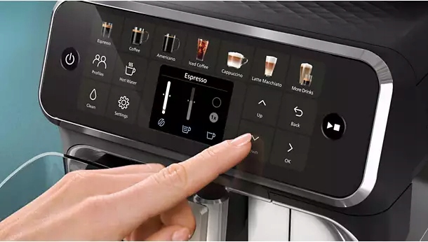Zbliżenie na dotykowy panel sterowania ekspresu Philips serii 5500. Dłoń wybiera ustawienia kawy na interaktywnym menu z kolorowymi ikonami napojów i centralnym wyświetlaczem parametrów parzenia. Srebrne wykończenie krawędzi panelu podkreśla nowoczesny design urządzenia.
