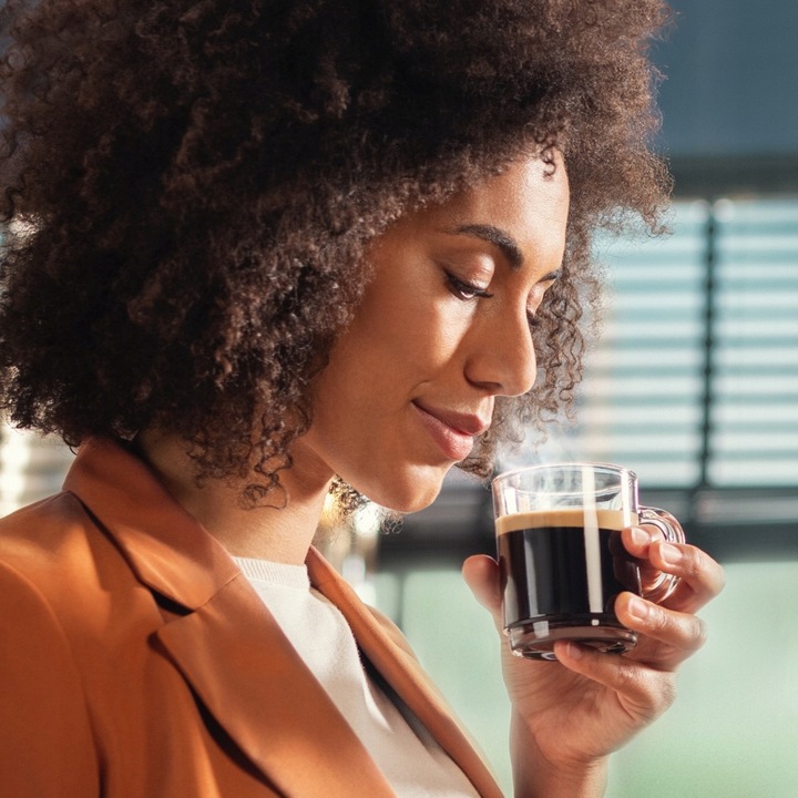 Kobieta delektująca się zapachem kawy espresso w przezroczystej filiżance. Zdjęcie w ciepłej tonacji przedstawia chwilę relaksu przy napoju z idealną cremą, przygotowanym w ekspresie Philips.