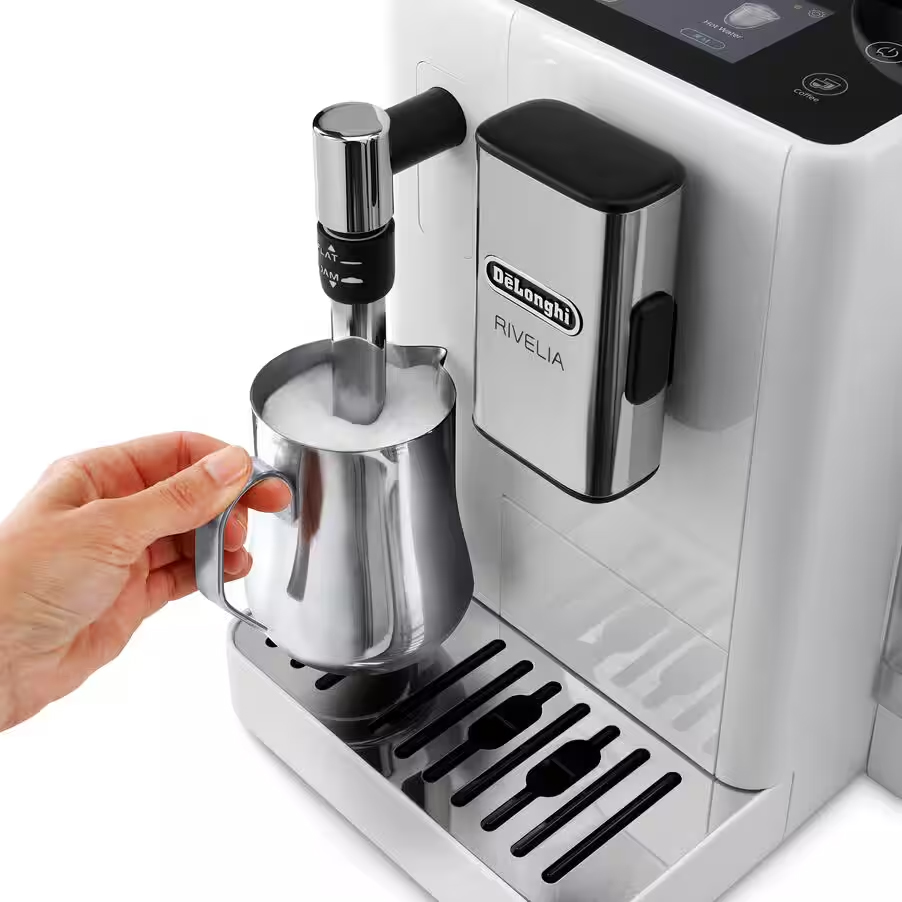 Zbliżenie na proces ręcznego spieniania mleka w stalowym dzbanku przy użyciu dyszy parowej w białym ekspresie DeLonghi Rivelia Arctic White. Dzbanek wypełniony białą pianą stoi na błyszczącej kratce ociekowej obok dozownika kawy z logo marki.