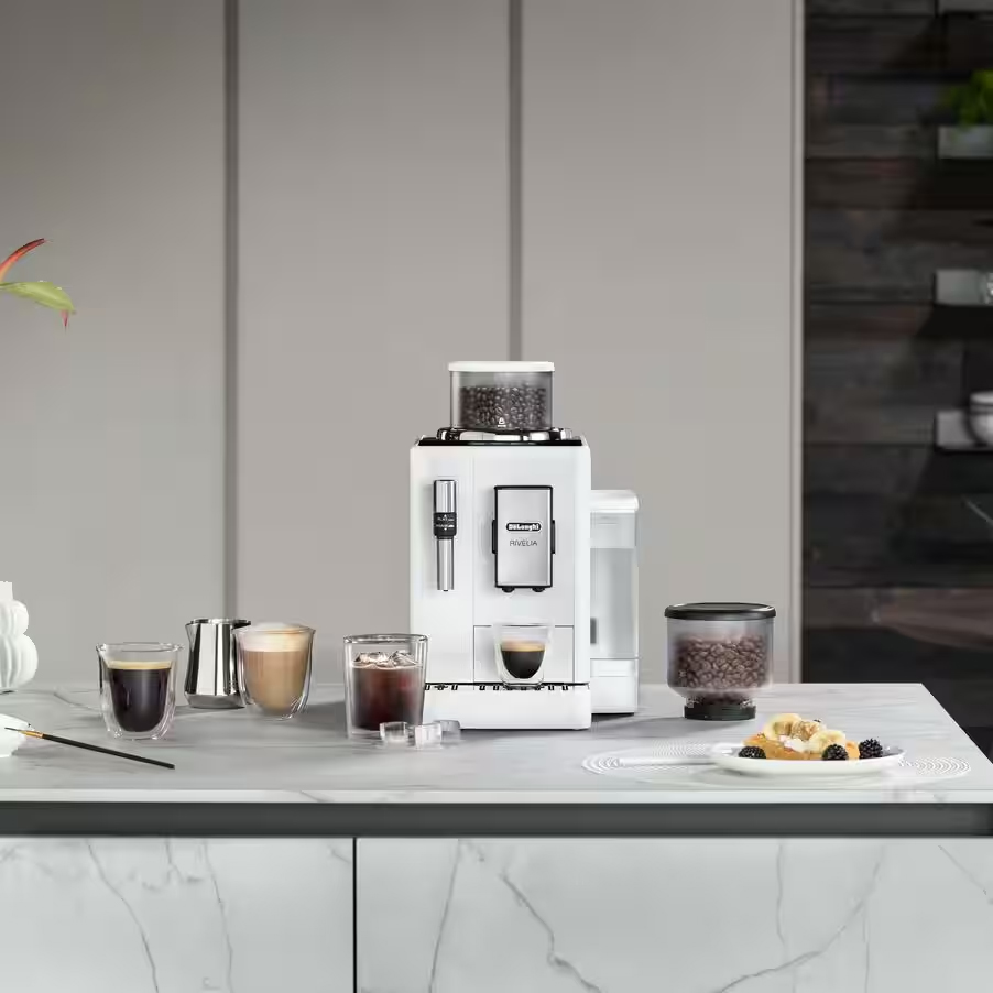 Biały ekspres DeLonghi Rivelia Arctic White wyeksponowany na jasnym, marmurowym blacie kuchennym. Wokół urządzenia ułożono różnorodne gotowe kawy, w tym mrożoną z kostkami lodu, deser na talerzyku oraz wymienny pojemnik na ziarna.
