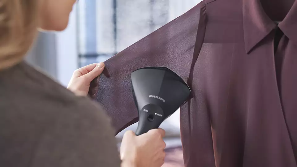 Zbliżenie na parownicę Philips GC800/80 podczas prasowania rękawa fioletowej koszuli. Widoczny tył urządzenia z wybranym trybem pracy oraz dłoń użytkownika napinająca materiał.