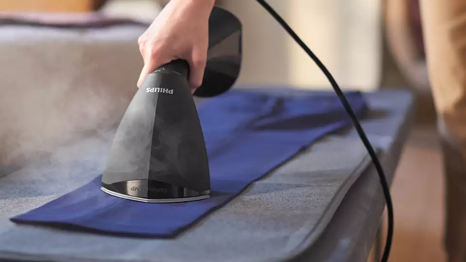 Zastosowanie ręcznego steamera Philips GC800/80 do wygładzania zagnieceń w poziomie na niebieskim materiale. Widoczna dłoń użytkownika dociskająca płytę parową z technologią OptimalTEMP do tkaniny na szarej macie, przy jednoczesnym wyrzucie delikatnej pary. Grafitowa obudowa parownicy posiada kontrastujące białe logo producenta.