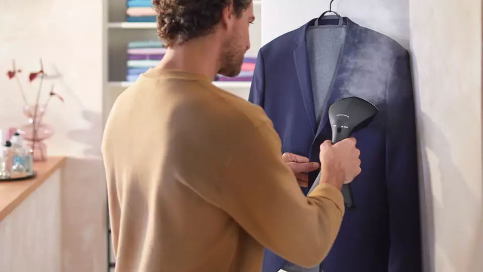 Mężczyzna odświeżający ciemnoniebieską marynarkę za pomocą ręcznego steamera Philips GC800/80. Widoczny intensywny wyrzut pary z urządzenia w kierunku tkaniny zawieszonej na wieszaku. Grafitowa obudowa parownicy z technologią OptimalTEMP pozwala na bezpieczne prasowanie w pionie, co zostało zaprezentowane w jasnym, domowym otoczeniu.