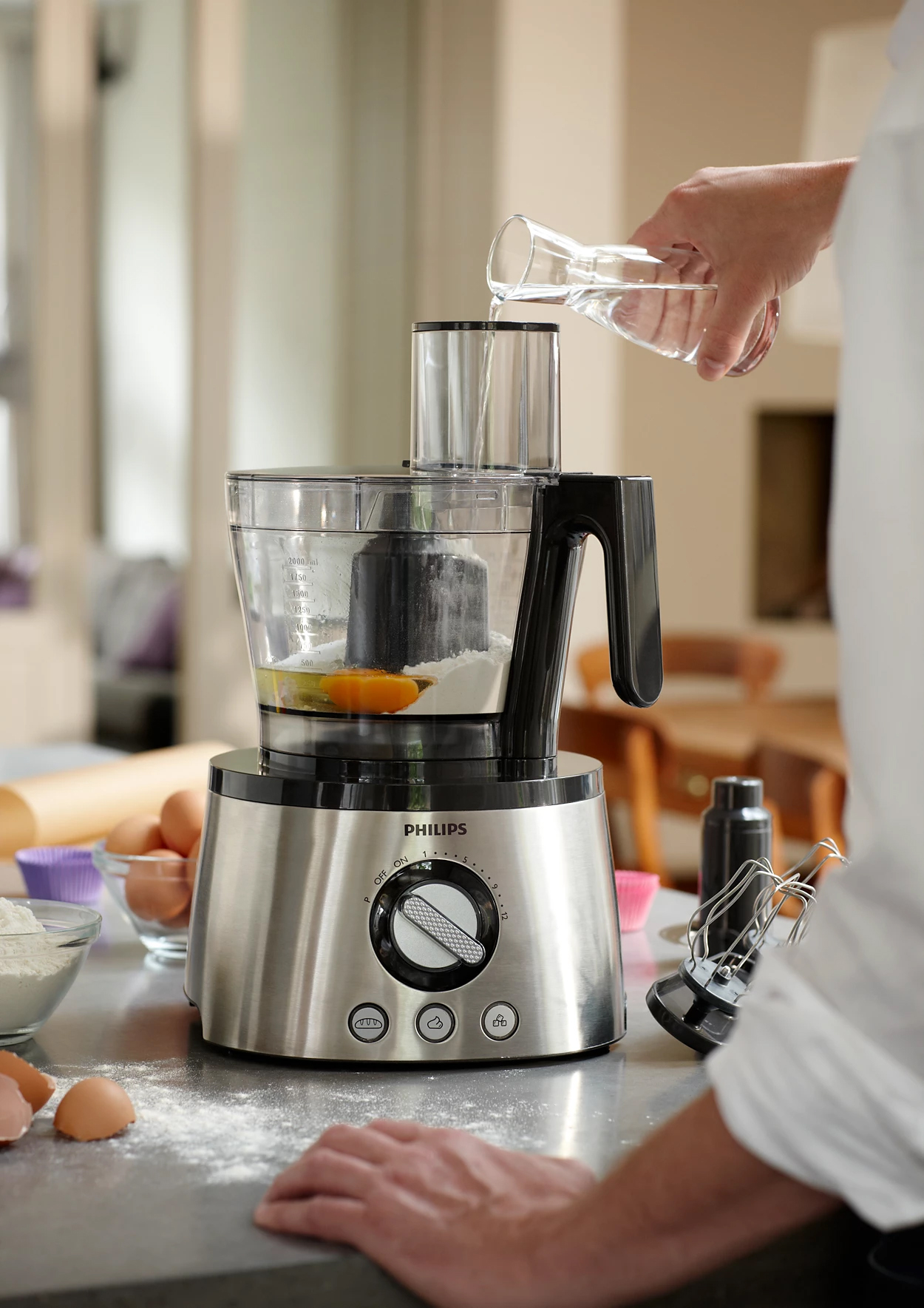 Robot kuchenny Philips HR7778/00 w trakcie pracy. Zdjęcie pokazuje proces dodawania wody do misy malaksera, w której znajdują się mąka i jajko. Urządzenie ze stali nierdzewnej stoi na blacie pośród składników do pieczenia i dodatkowych przystawek z zestawu.