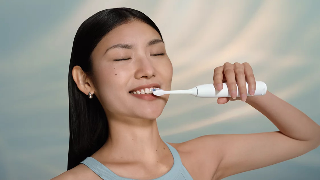 Kobieta o azjatyckiej urodzie myje zęby białą szczoteczką soniczną Philips Sonicare. Trzyma włączone urządzenie przy zębach prawą ręką, ma zamknięte oczy i uśmiecha się. Tło jest jasne i rozmyte.