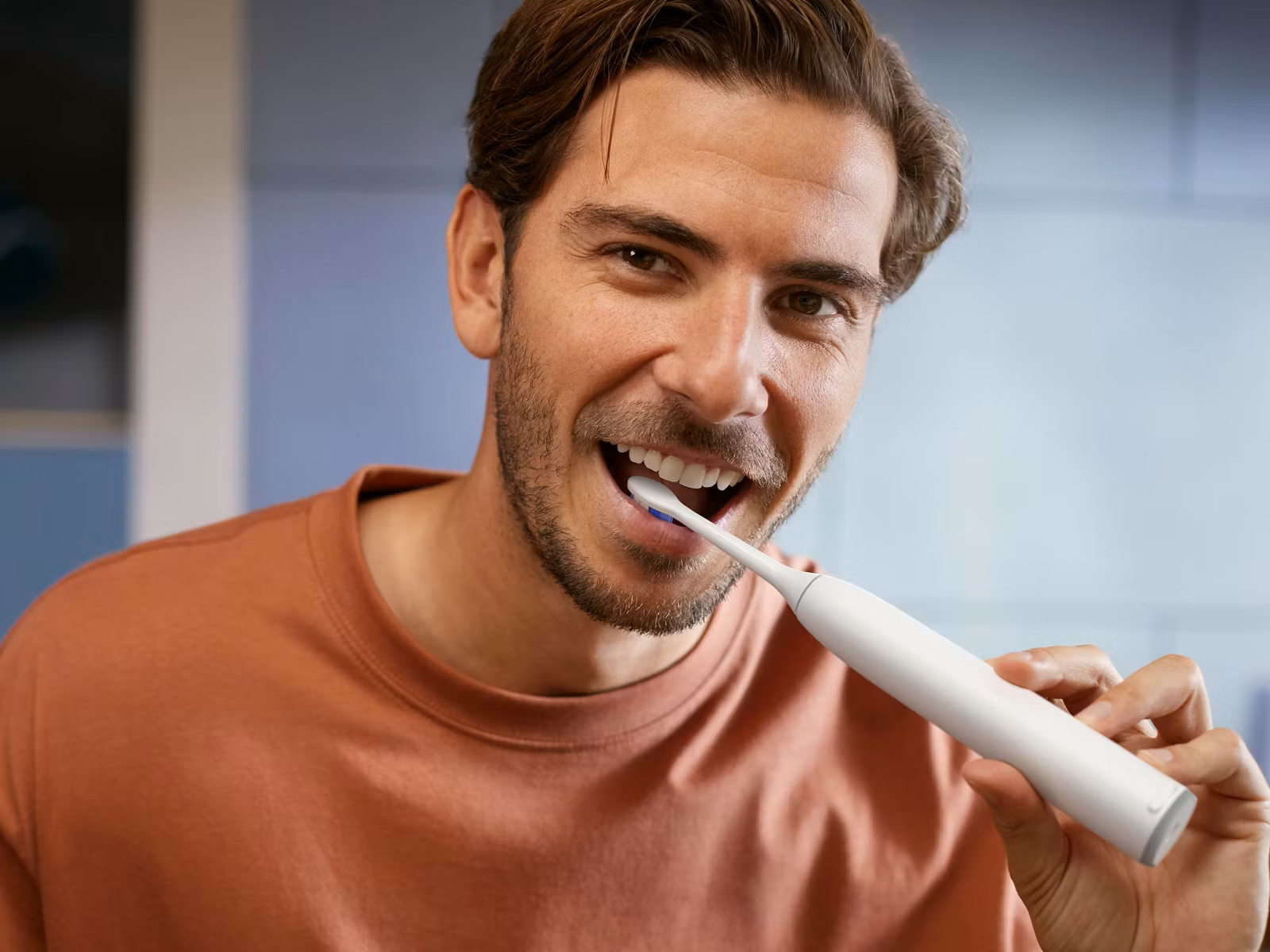 Uśmiechnięty mężczyzna myje zęby białą szczoteczką soniczną Philips Sonicare 6500. Szczoteczka jest włożona do jego ust, demonstrując użycie produktu. Tło stanowi łazienka.