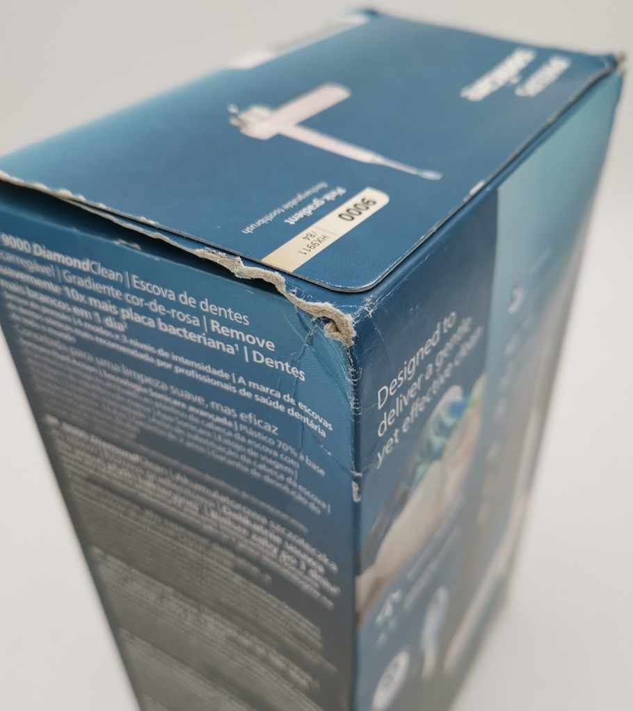 Zbliżenie na uszkodzony, zgnieciony narożnik niebieskiego pudełka szczoteczki Philips Sonicare 9000 DiamondClean HX9911/84. Na opakowaniu widoczne są napisy techniczne oraz naklejka z numerem modelu na górnej klapie.