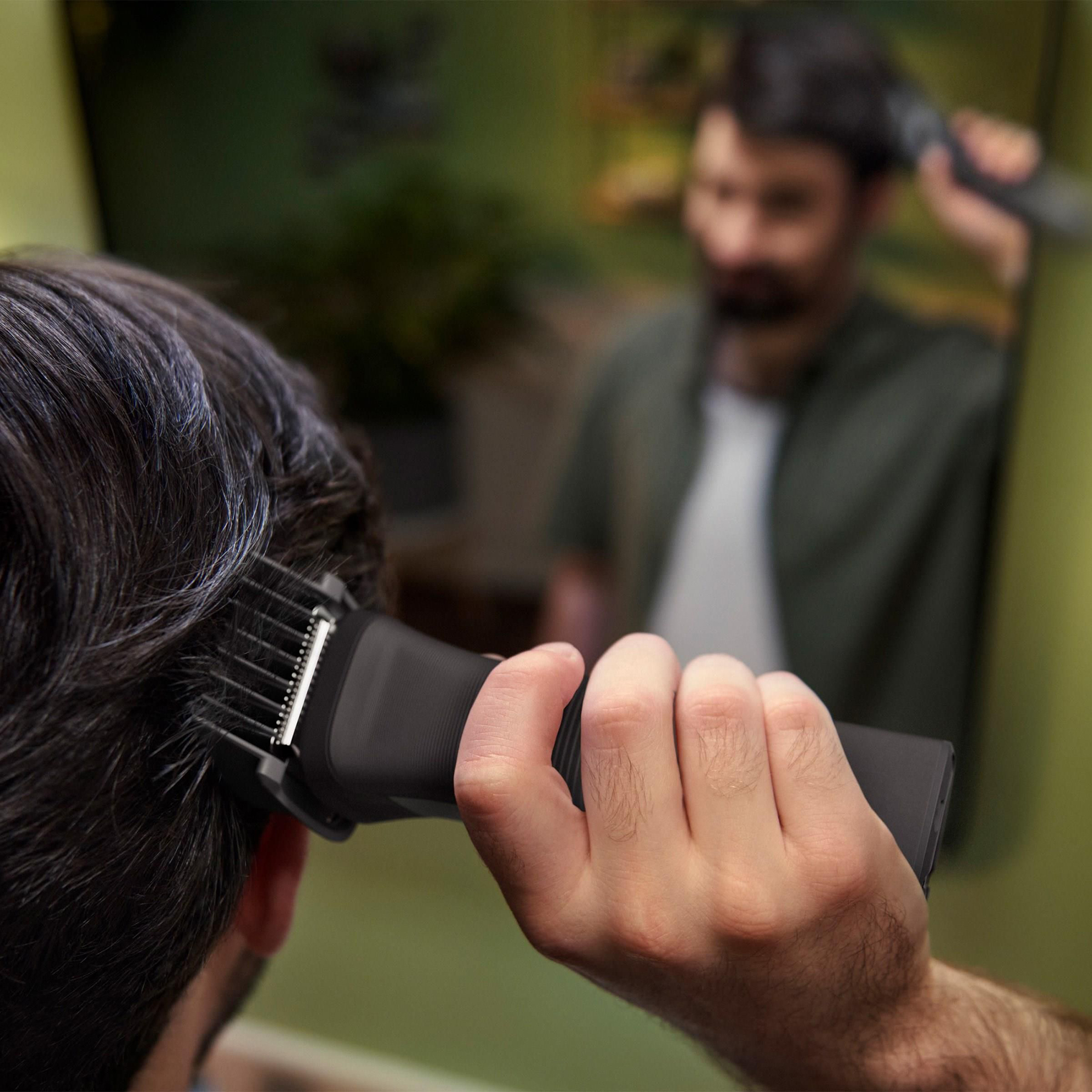 Zbliżenie na męską dłoń trzymającą czarny trymer Philips Multigroom MG7940/15 z nasadką, który strzyże włosy z tyłu głowy. W tle, w rozmyciu, widać odbicie w lustrze mężczyzny w domowej łazience, co demonstruje samodzielne użycie urządzenia.