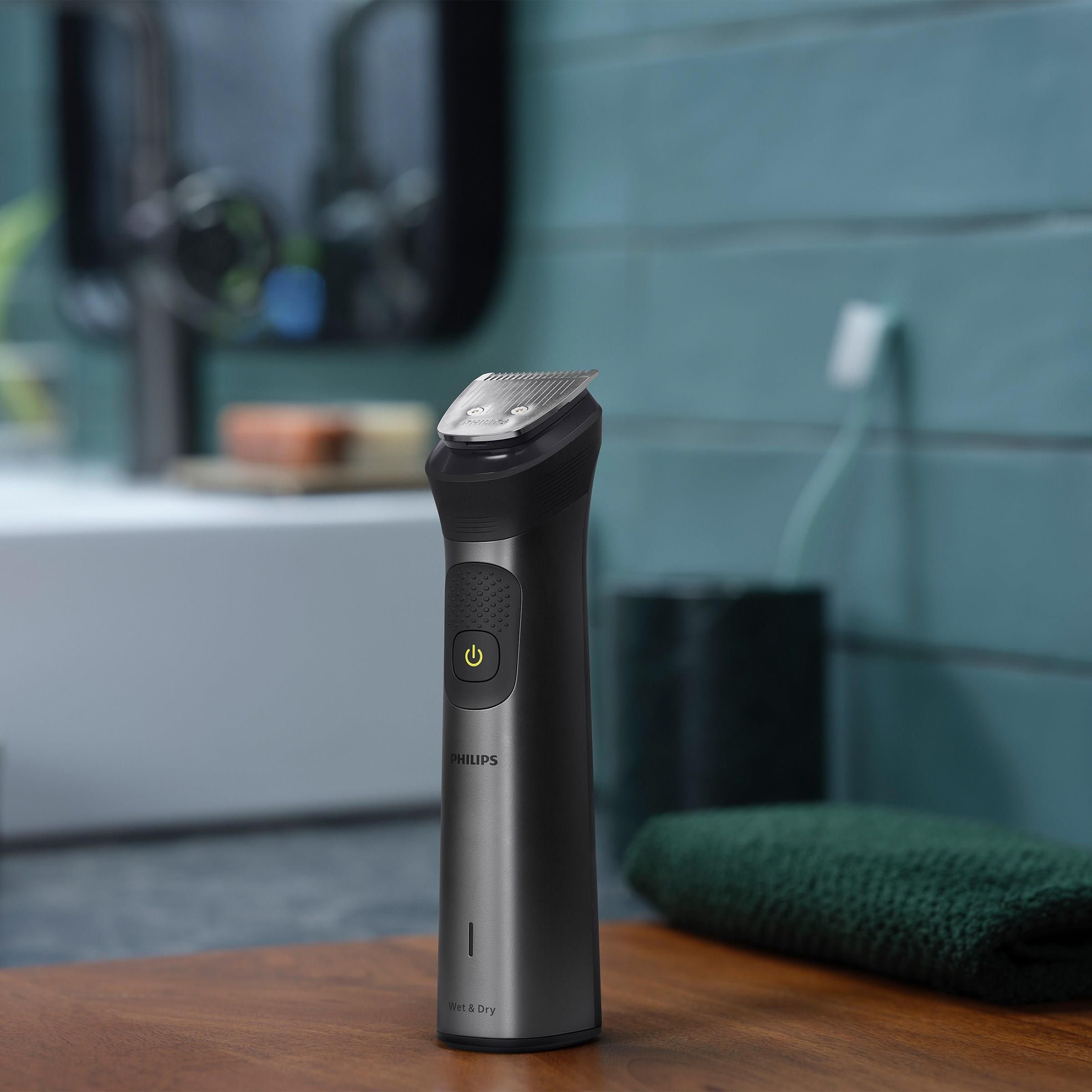 Trymer Philips Multigroom MG7940/15 ustawiony na drewnianym blacie w łazience. Nowoczesny design urządzenia w kolorze ciemnego metalu kontrastuje z turkusowym tłem ściany i zielonym ręcznikiem, podkreślając domowy kontekst użytkowania.