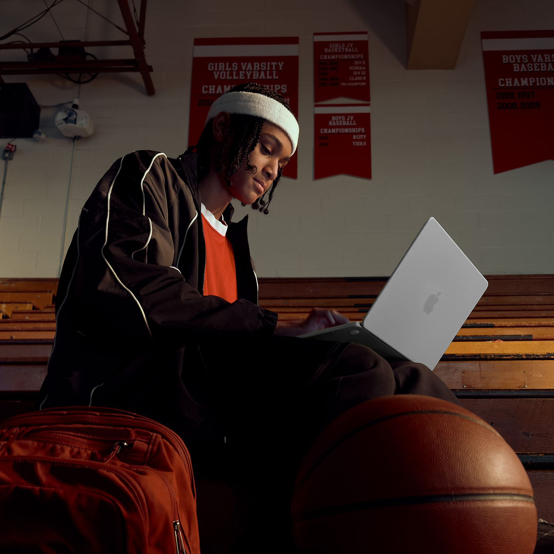 Srebrny MacBook Neo 2026 używany przez młodego sportowca na trybunach hali gimnastycznej. Obok widoczna piłka do koszykówki i plecak. Dynamiczne ujęcie łączące naukę lub pracę z pasją do sportu.