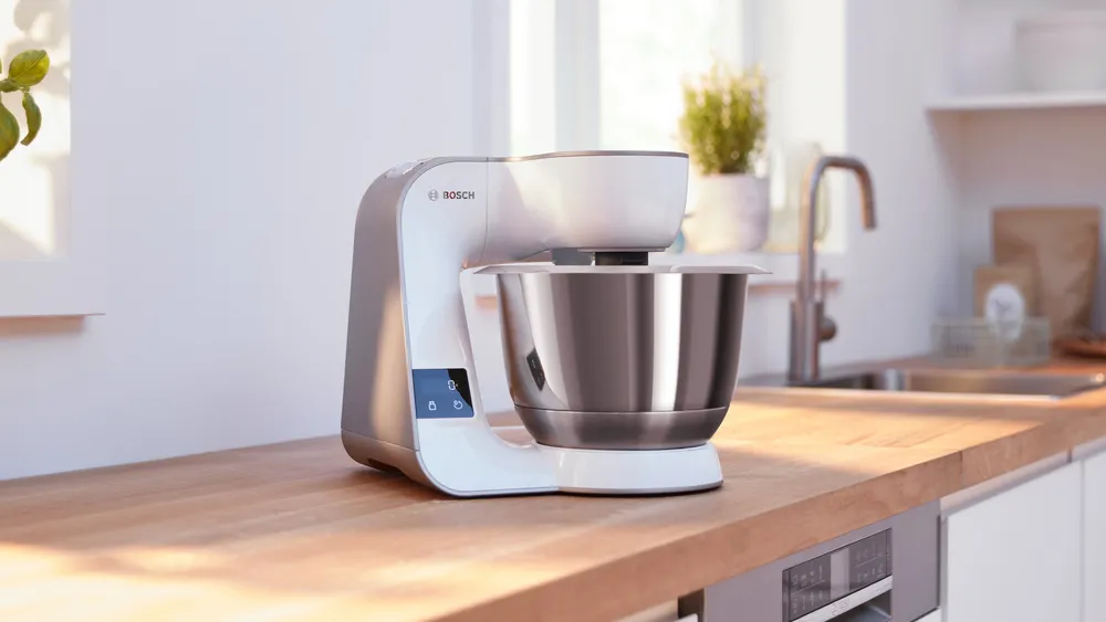 Robot kuchenny Bosch MUM 5 MUM5XW20 ze stalową misą stoi na drewnianym blacie w jasnej kuchni, widok z boku. Na włączonym wyświetlaczu widnieje cyfra 0, a w tle widać okno, zlew oraz szafki kuchenne.
