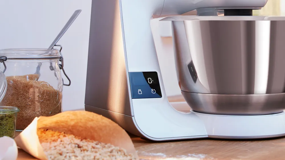 Zbliżenie na robot kuchenny Bosch MUM 5 MUM5XW20 ze stalową misą na drewnianym blacie. Na małym ekranie widać wartość 0 g, a obok stoją słoik z brązowym cukrem oraz torba z produktami sypkimi.