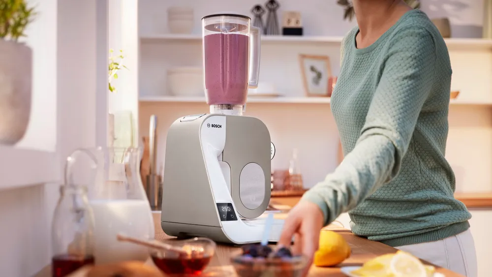 Robot kuchenny Bosch MUM 5 MUM5XW20 z przezroczystym blenderem kielichowym pełnym fioletowego koktajlu na drewnianym blacie w kuchni. Kobieta w zielonej bluzce sięga po miseczkę z owocami stojącą obok urządzenia.