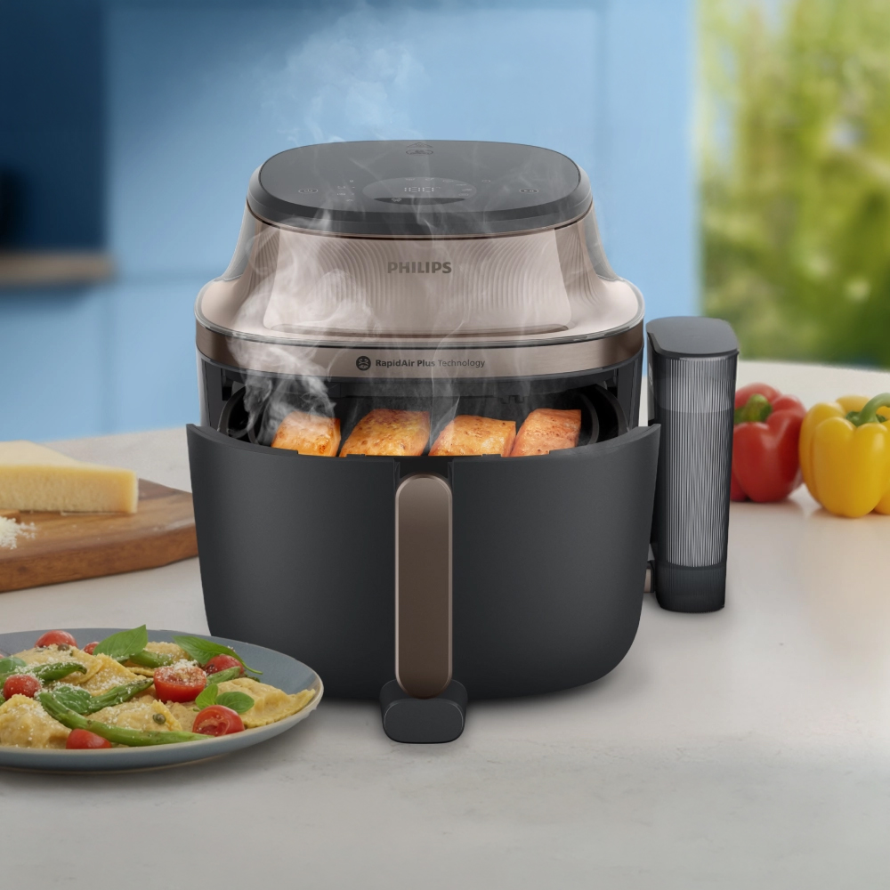 Frytkownica Philips Airfryer NA547/07 z uchyloną szufladą, z której unosi się para nad upieczonym łososiem. Urządzenie stoi na blacie kuchennym obok talerza z ravioli i papryk. Z boku przymocowano pojemnik na wodę.