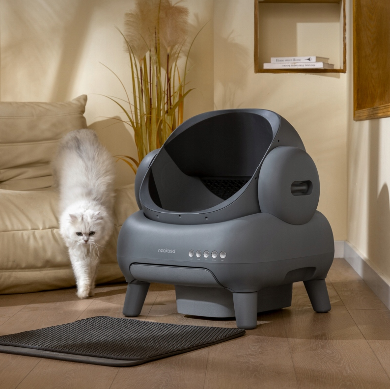 Szara kuweta Neakasa M1 w domowym wnętrzu z widoczną przed nią matą na żwirek. Biały kot zbliża się do urządzenia, co pokazuje codzienne użytkowanie produktu. Nowoczesny wygląd kuwety komponuje się z jasnym salonem i beżową sofą.