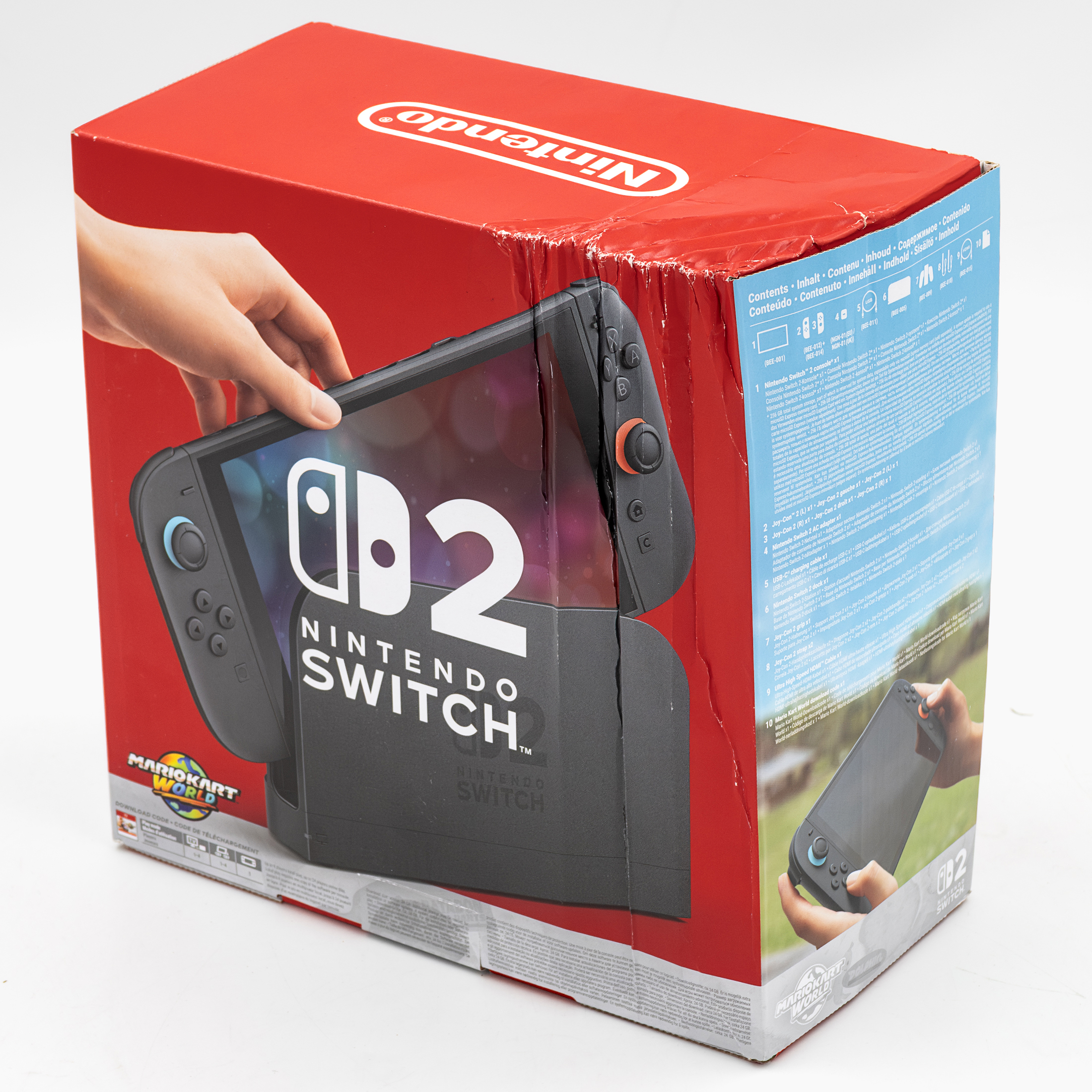 Pudełko od konsoli Nintendo Switch 2 z zestawu Mario Kart World, widok pod kątem na białym tle. Kartonowe opakowanie jest wyraźnie uszkodzone, pogniecione i nadszarpnięte wzdłuż górnej oraz bocznej krawędzi. Na froncie widoczna jest grafika urządzenia, a na niebieskim boku specyfikacja.