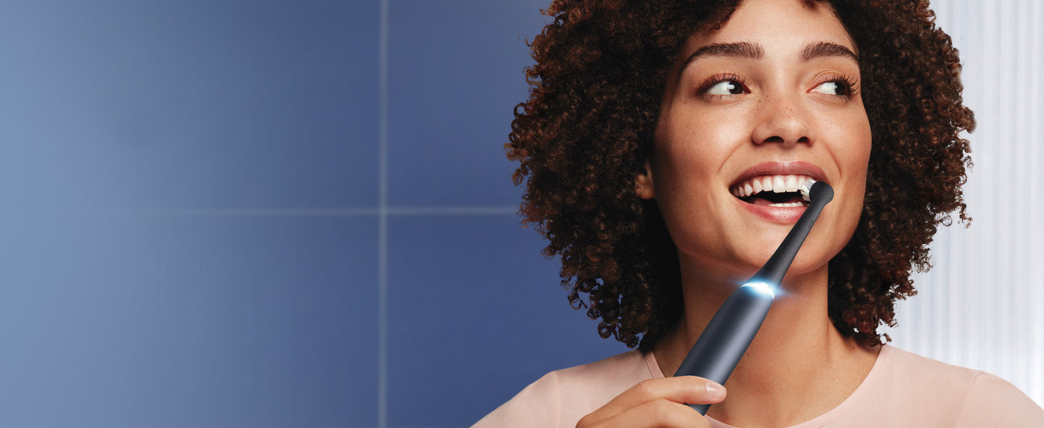 Uśmiechnięta kobieta ze szczoteczką elektryczną Oral-B iO w ustach, w łazience z niebieską ścianą.