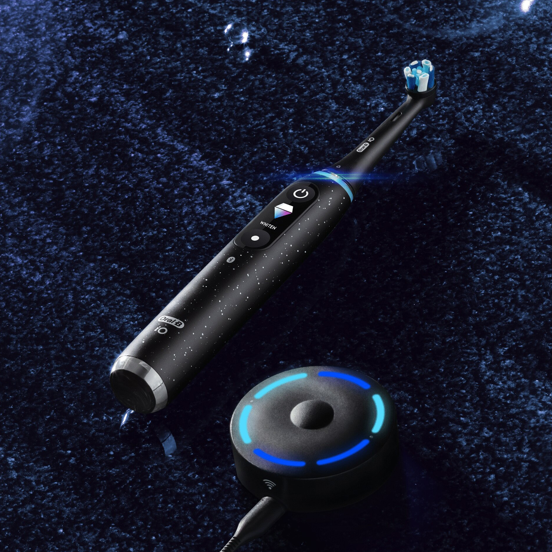 Czarna szczoteczka magnetyczna Oral-B iO Series 10 z włączonym wyświetlaczem pokazującym tryb WHITEN. Szczoteczka leży obok okrągłej, czarnej stacji ładującej z niebieskim podświetleniem LED, na ciemnym tle.
