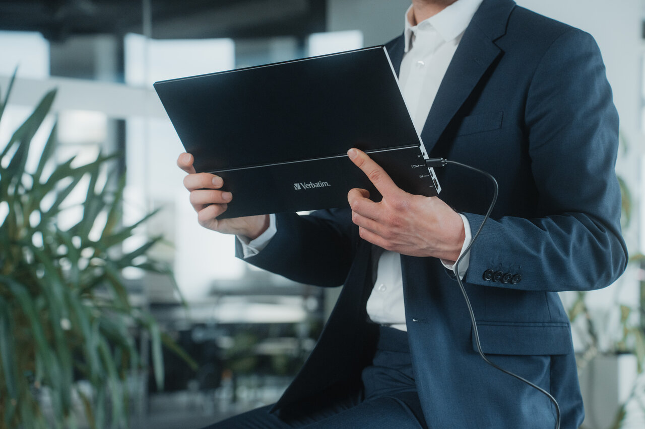 Mężczyzna w garniturze trzyma w dłoniach złożony, czarny przenośny monitor Verbatim. Widoczne jest logo Verbatim na obudowie oraz podłączony czarny przewód. Scena rozgrywa się w nowoczesnym biurze.