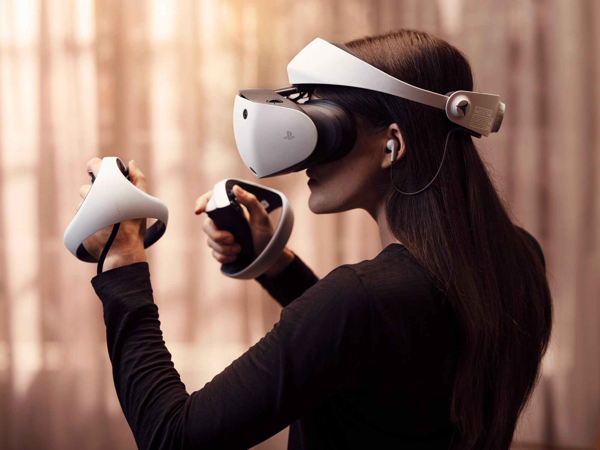 Kobieta z profilu testuje gogle wirtualnej rzeczywistości Sony PlayStation VR2 i trzyma w dłoniach dwa kontrolery Sense. Ma na sobie czarną bluzkę z długim rękawem oraz białą słuchawkę douszną. Urządzenie oraz zaokrąglone kontrolery odznaczają się jasną, białą kolorystyką na rozmytym, ciepłym tle.