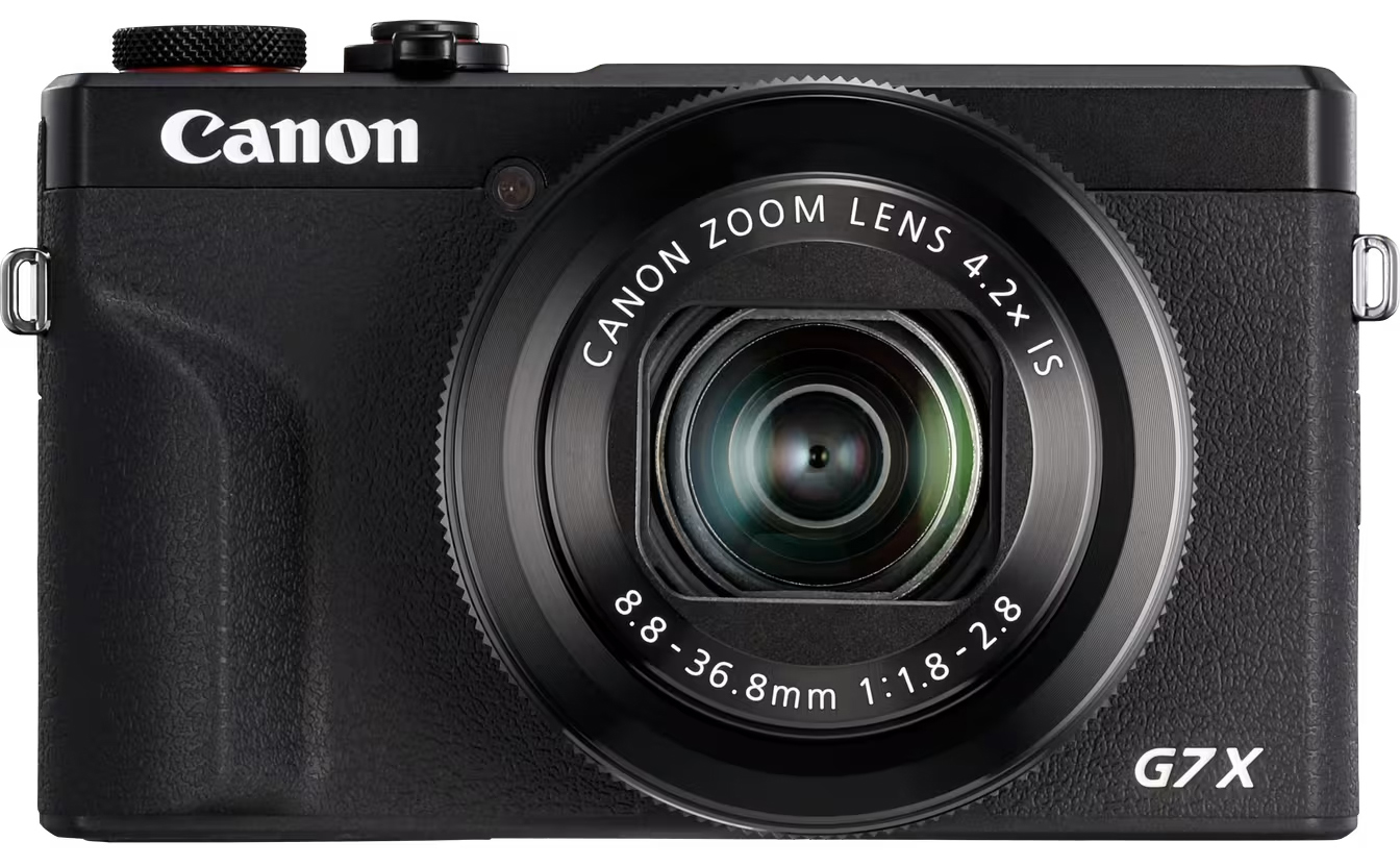 Czarny aparat kompaktowy Canon PowerShot G7X Mark III na białym tle, widok od przodu. Korpus ma chropowatą strukturę z białym logo CANON oraz wyraźnym oznaczeniem modelu G7X. Centralne miejsce zajmuje duży obiektyw zmiennoogniskowy z widocznymi parametrami optycznymi oraz czerwonym akcentem przy górnym pokrętle.